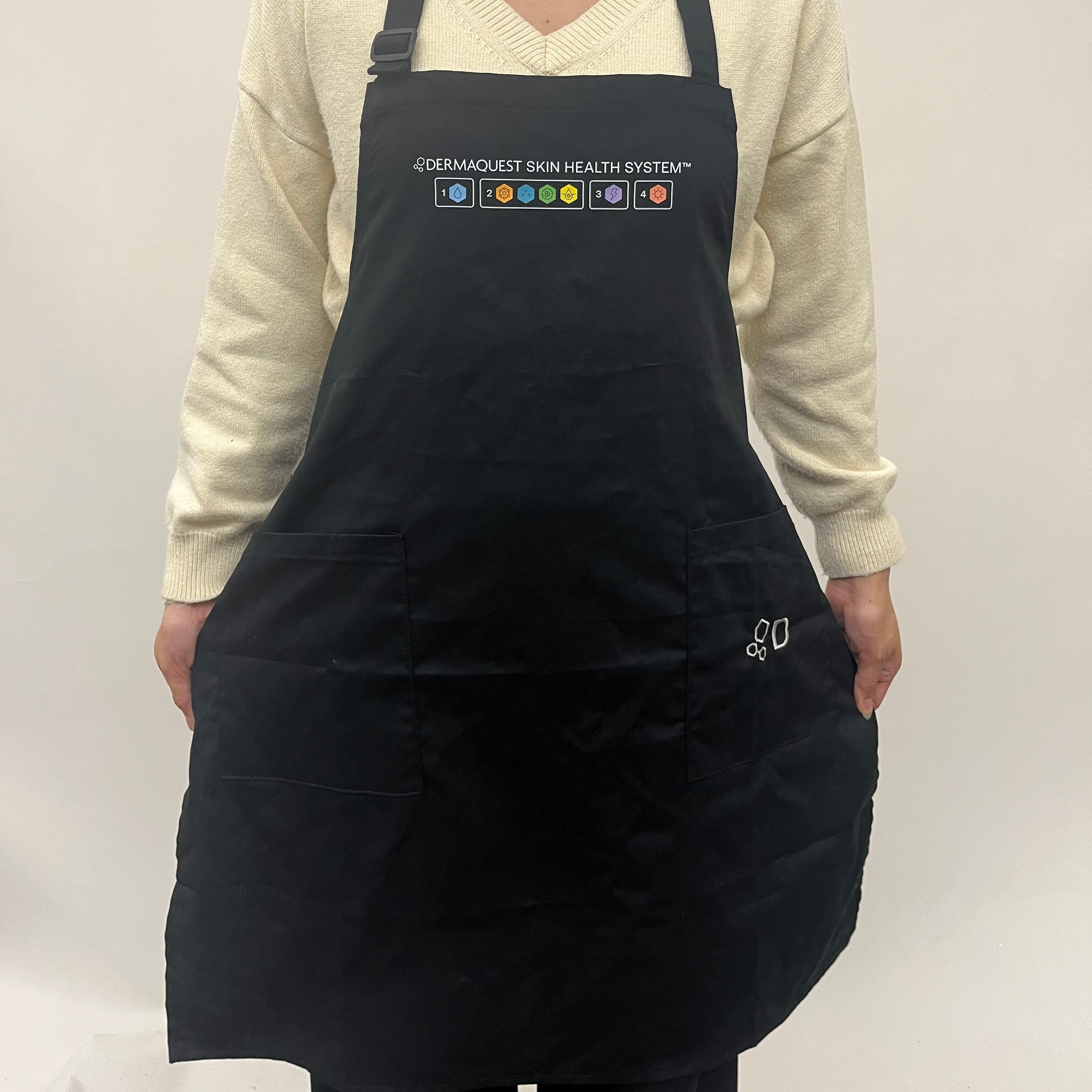 A person wearing a black apron over a cream-colored sweater, standing against a plain light background. The apron features a white printed logo and text at the chest reading 'DERMAQUANT SKIN HEALTH FOUNDATION' with colorful icons, and a small white monogram on the lower right pocket.