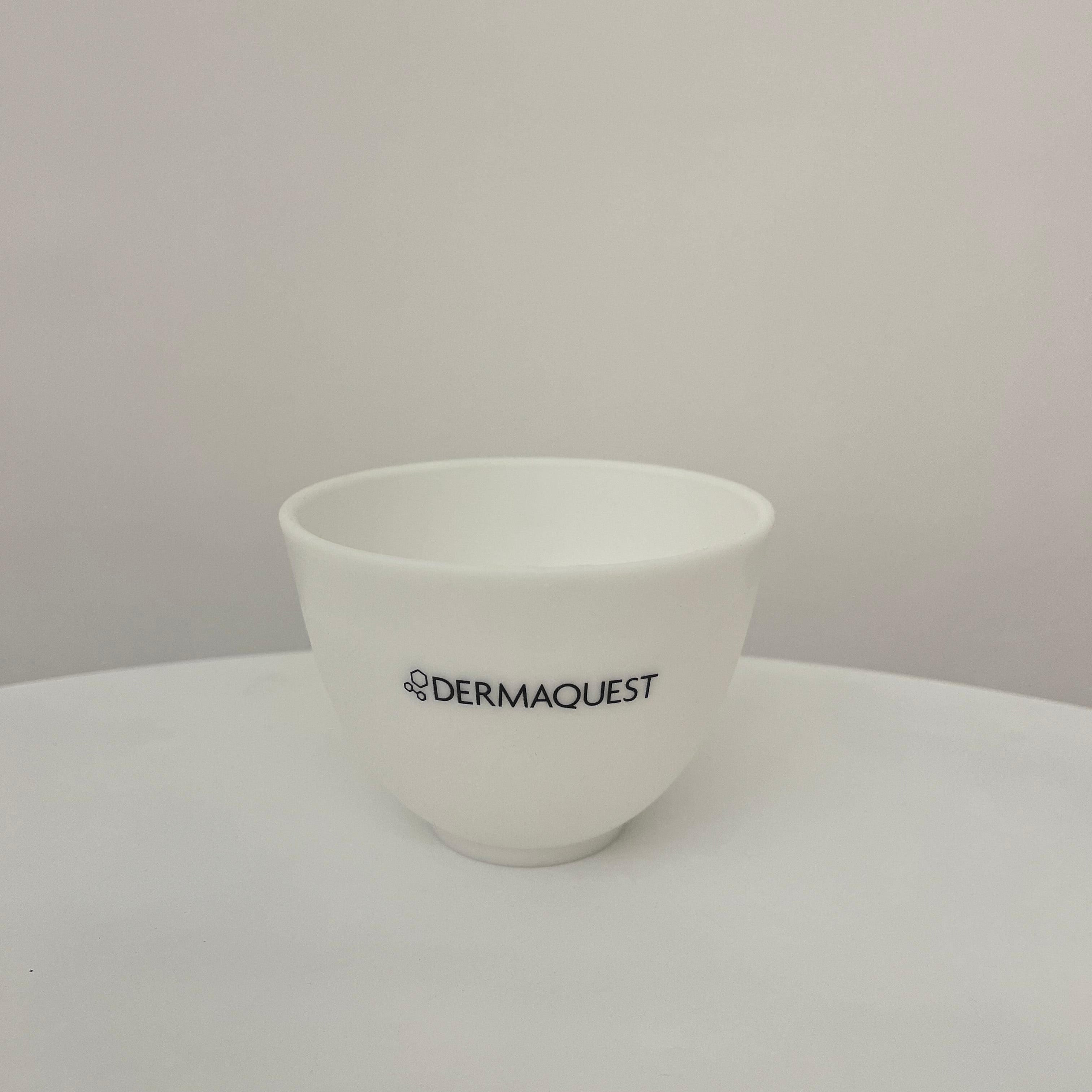 A white ceramic cosmetic bowl with the black printed brand name 'DERMAQUEST' on the front, sitting on a white surface against a neutral light gray background.