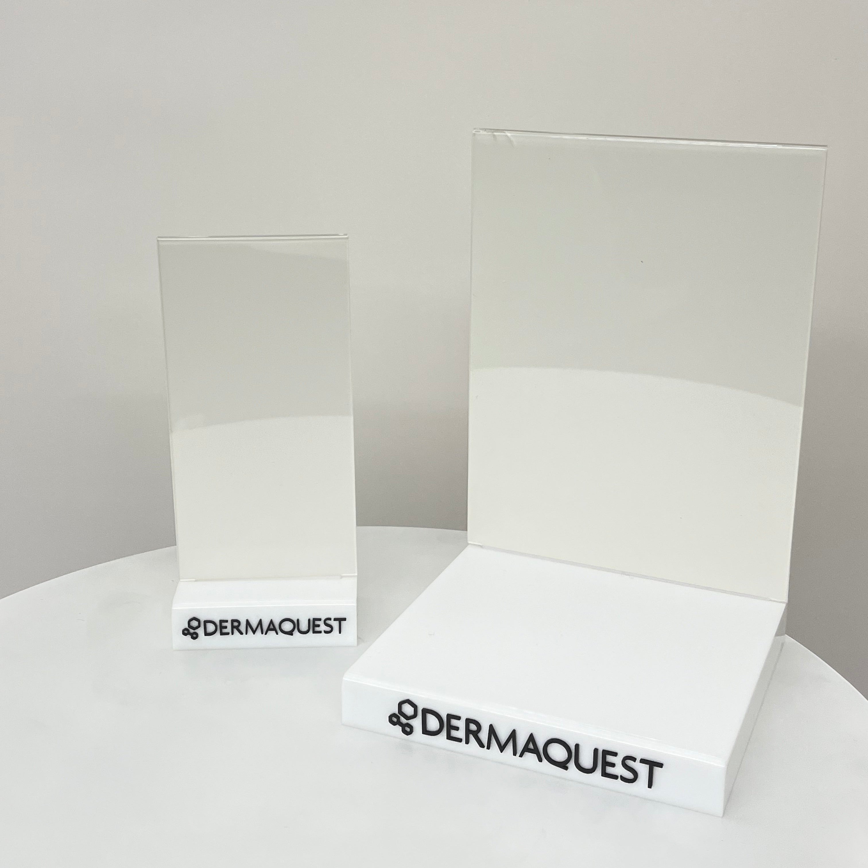 Two clear acrylic display blocks with white bases printed with the black DERMAQUEST logo, displayed on a white surface against a light gray background.